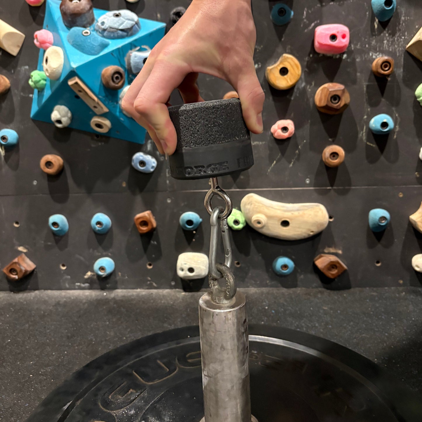 Hand holding a weight on a climbing hold with a weight attached, against a colorful climbing wall.